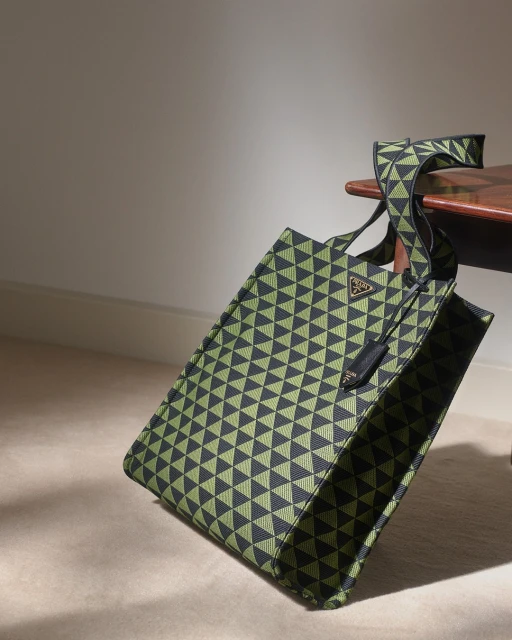 Studio shot of a geometric patterned Prada tote bag in green and black resting on a dark wooden table in front of a neutral wall.