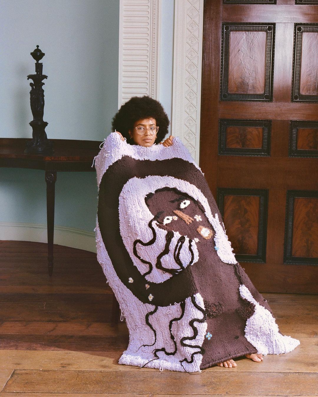 A person with an afro and glasses peeks from behind a large, colorful, patterned rug, posing in a light-blue-walled room with wooden floors.
