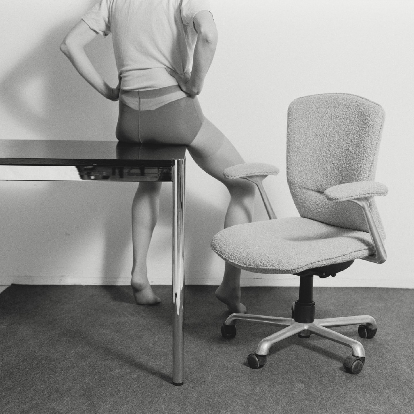 Monochrome shot of a person sitting on a glossy black desk, legs visible, with an office chair nearby, set against a plain white background.