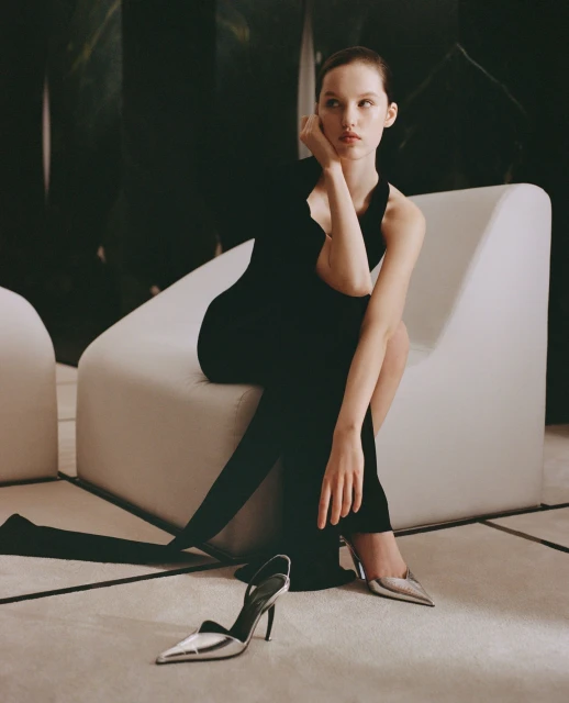 Elegant woman in a black dress poses seated on a white chair with silver high-heeled shoes on the floor, creating a high fashion aesthetic.