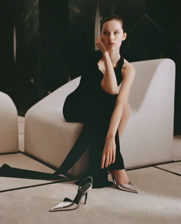 Elegant woman in a black dress poses seated on a white chair with silver high-heeled shoes on the floor, creating a high fashion aesthetic.