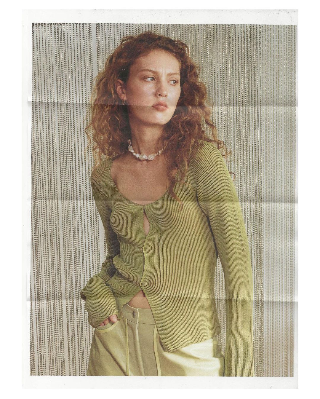 Woman with curly hair wearing a sage green cardigan and light yellow pants against a neutral striped background in an indoor setting.