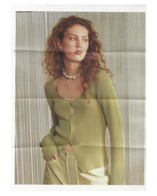 Woman with curly hair wearing a sage green cardigan and light yellow pants against a neutral striped background in an indoor setting.