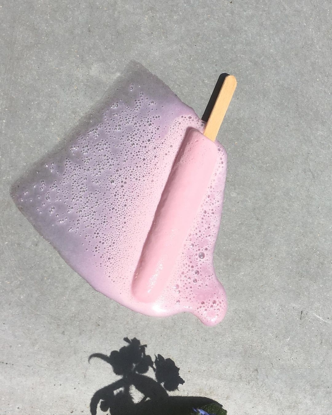 A melted pink popsicle with a wooden stick sits on a gray surface in bright, natural light, casting a shadow of a flower.