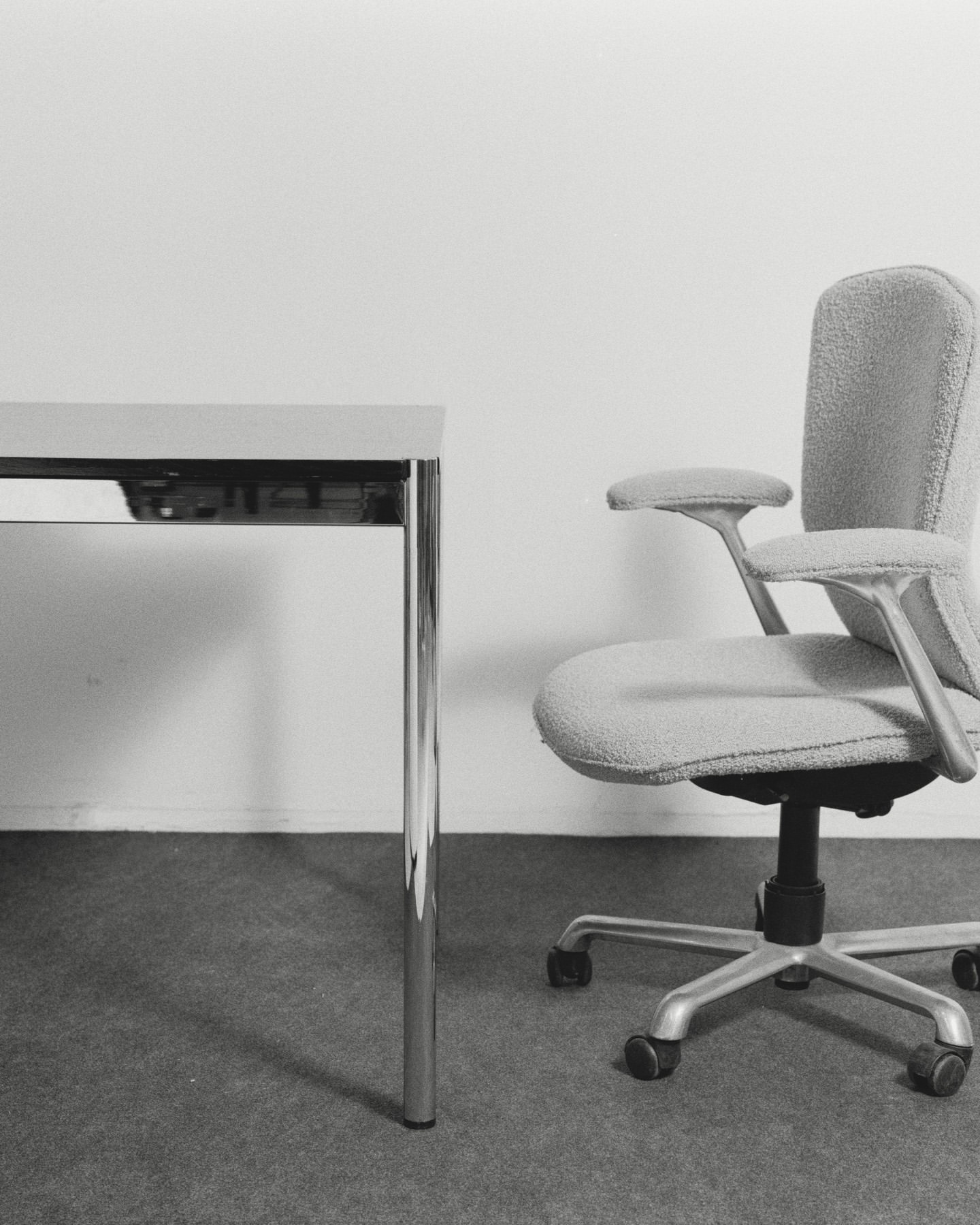 Office interior featuring a gray upholstered office chair with armrests next to a gray table, set against a plain white wall.