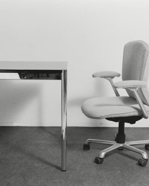 Office interior featuring a gray upholstered office chair with armrests next to a gray table, set against a plain white wall.