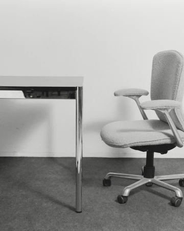 Office interior featuring a gray upholstered office chair with armrests next to a gray table, set against a plain white wall.