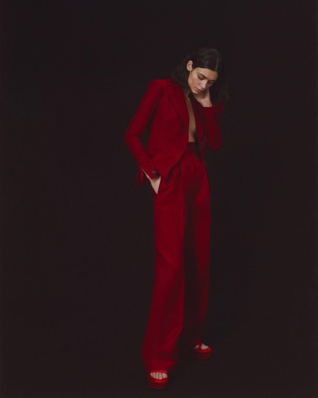 Elegant adult woman in a red suit posing against a black backdrop, showcasing fashion and style with a confident stance and modern attire.