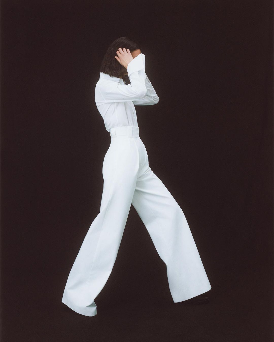 A person in a white long-sleeved shirt and high-waisted, wide-leg trousers poses on a dark background in a fashion studio shot.