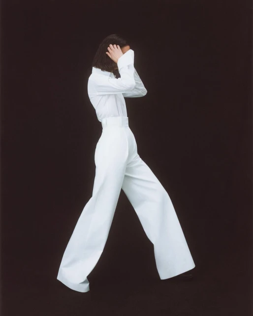 A person in a white long-sleeved shirt and high-waisted, wide-leg trousers poses on a dark background in a fashion studio shot.