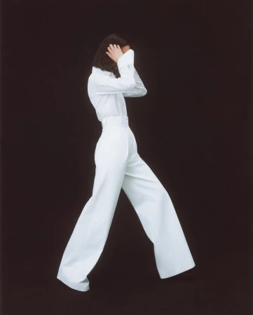 A person in a white long-sleeved shirt and high-waisted, wide-leg trousers poses on a dark background in a fashion studio shot.