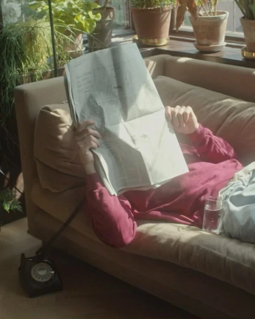 Adult reading newspaper on couch next to vintage rotary phone and glass of water in sunlit room with potted plants.
