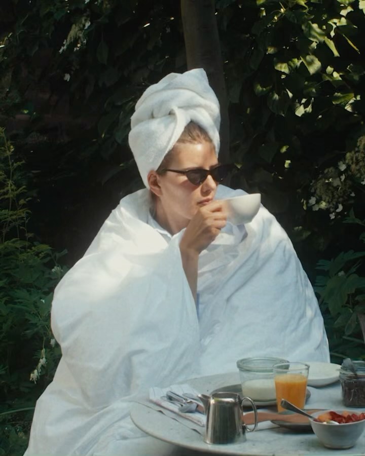 Adult female drinks from a teacup, wearing sunglasses and a white towel wrapped around her head and shoulders in an outdoor setting.