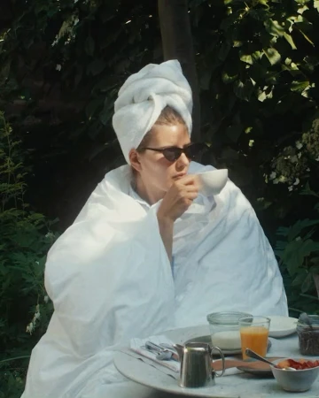 Adult female drinks from a teacup, wearing sunglasses and a white towel wrapped around her head and shoulders in an outdoor setting.