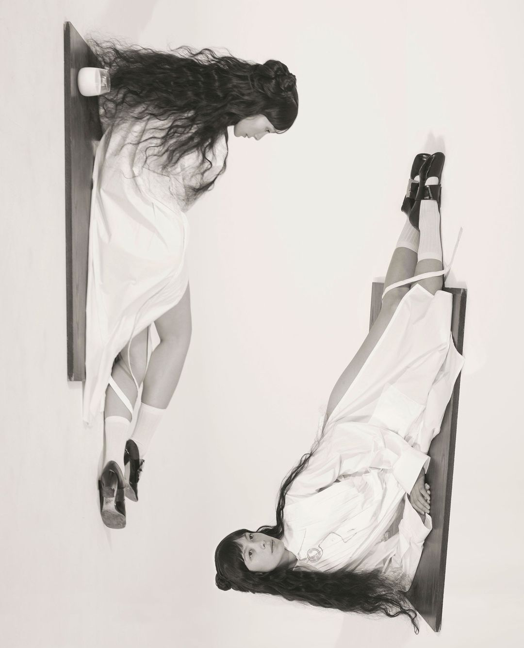 Black and white portrait features two women in white dresses, one looking down and the other looking up, floating on wooden shelves.