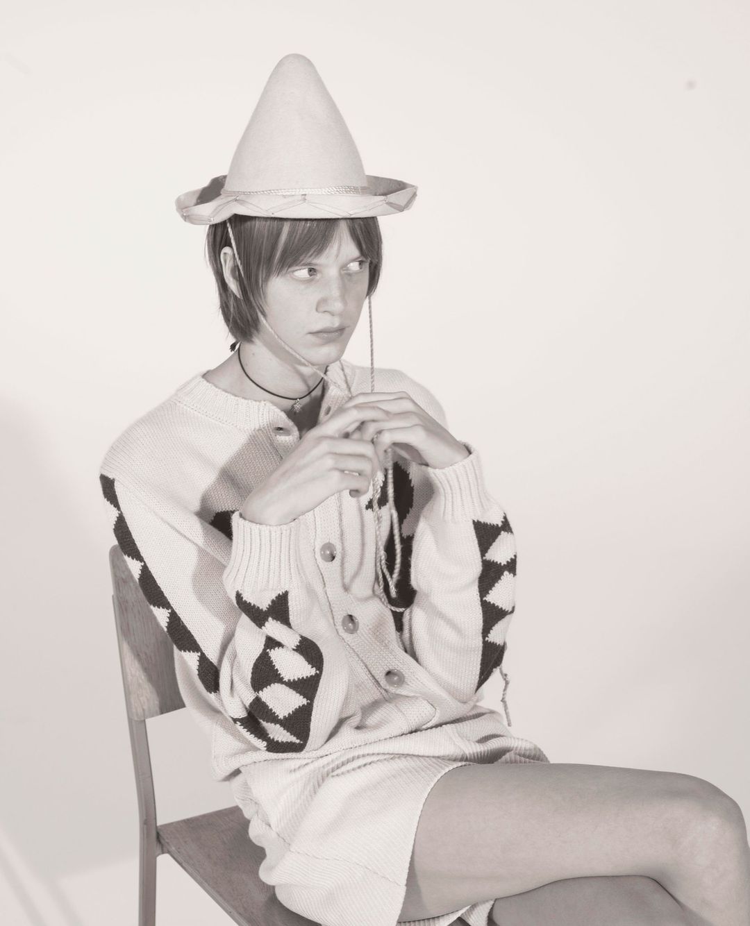 Studio shot of a person sitting on a chair wearing a hat, cardigan, and skirt, with a light grey background.