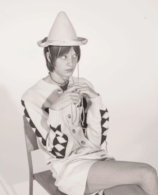 Studio shot of a person sitting on a chair wearing a hat, cardigan, and skirt, with a light grey background.
