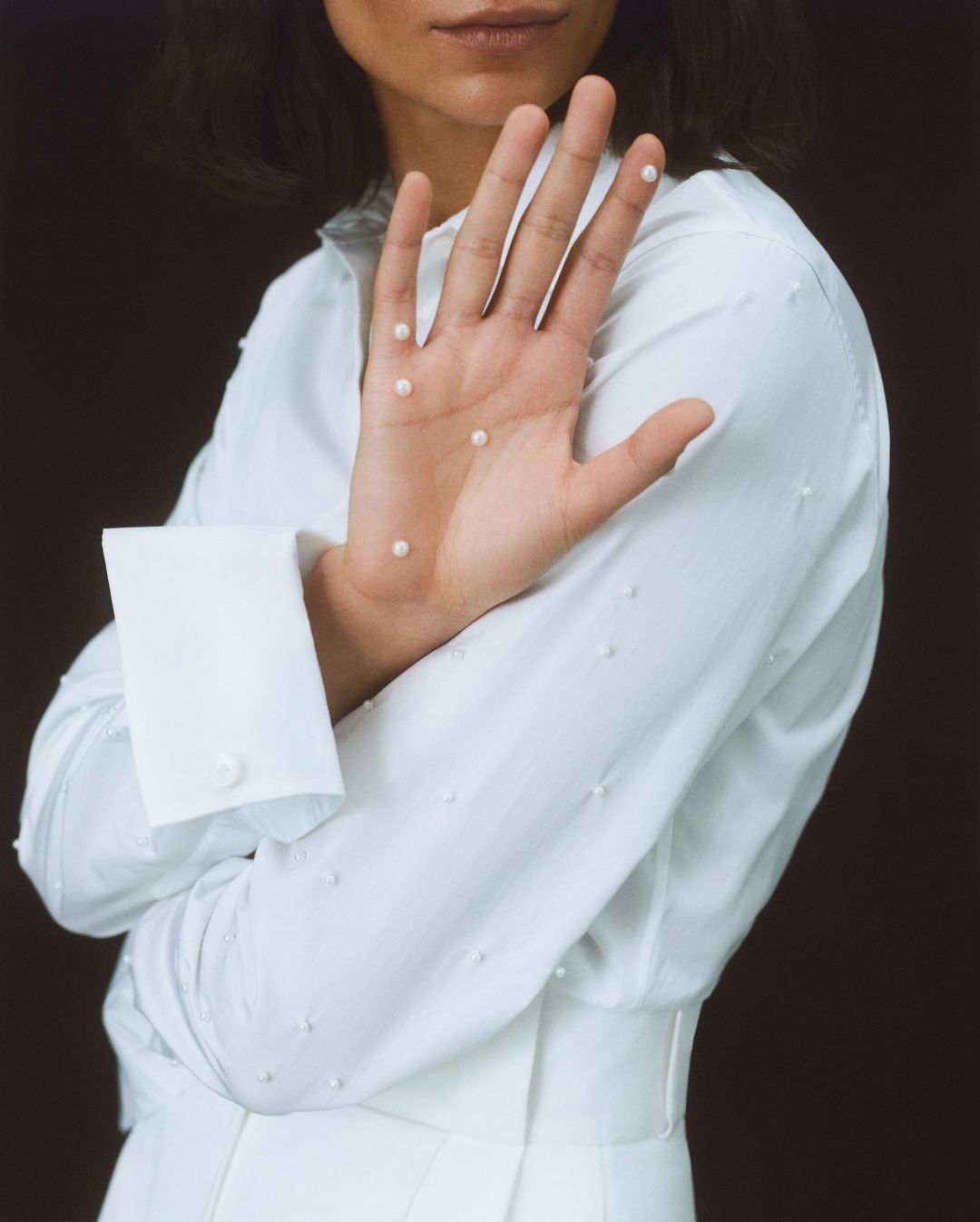 Woman in a white outfit with tiny pearls on her hand, posing against a dark backdrop, in a beauty and fashion-focused composition.