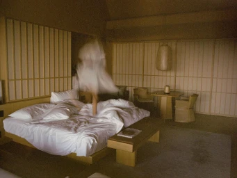 A blurred figure in a white dress jumping on a rumpled bed inside a wood-paneled hotel room photographed by Polly Brown.