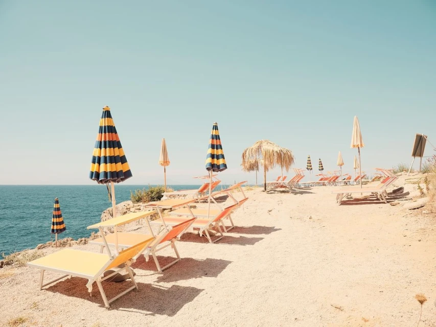 Beachside scene featuring lounge chairs and striped umbrellas under a clear sky, capturing a serene coastal vacation vibe.