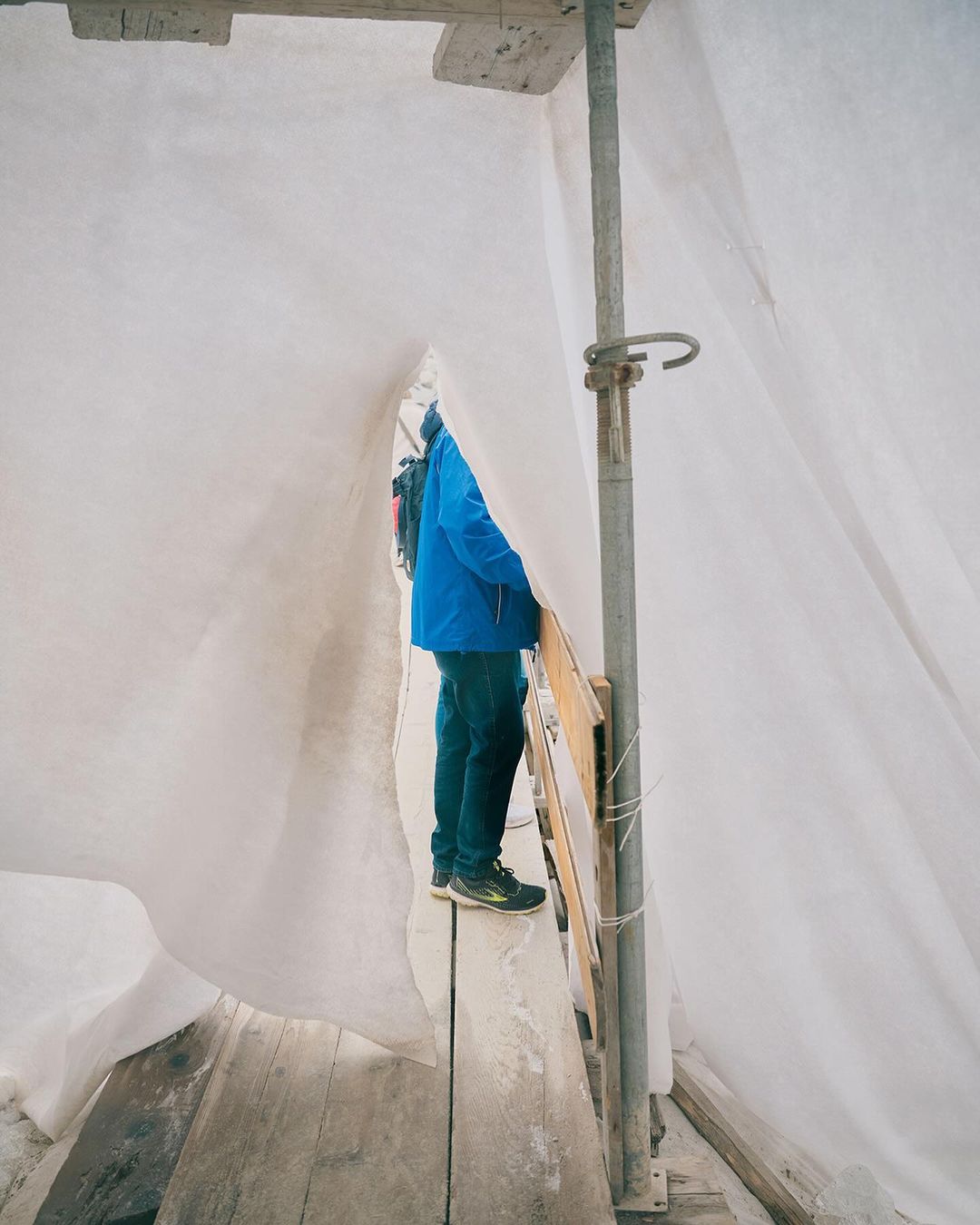 Person in blue jacket stands on wooden planks amidst scaffolding covered with white sheeting, construction or renovation in progress.