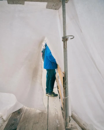 Person in blue jacket stands on wooden planks amidst scaffolding covered with white sheeting, construction or renovation in progress.