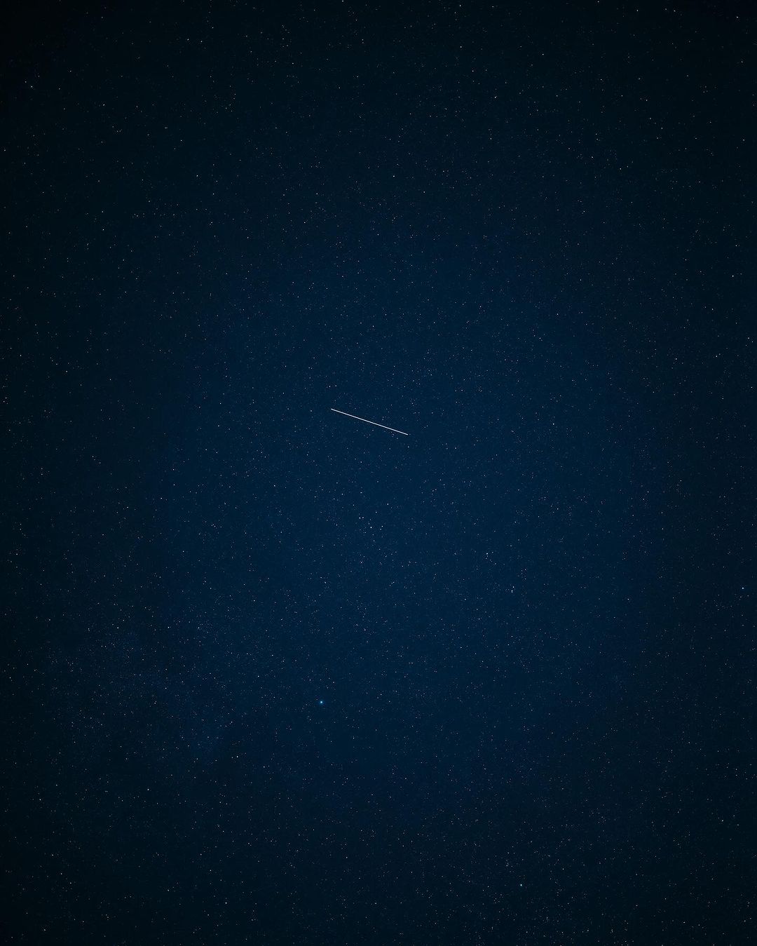 Night sky with numerous stars and a linear streak across the frame, possibly a meteor or satellite trail, under dark conditions.
