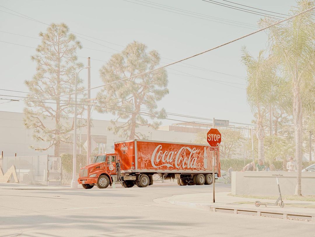 Red Coca-Cola delivery truck on urban street corner with faded light, trees, buildings, stop sign, and scooter in background.