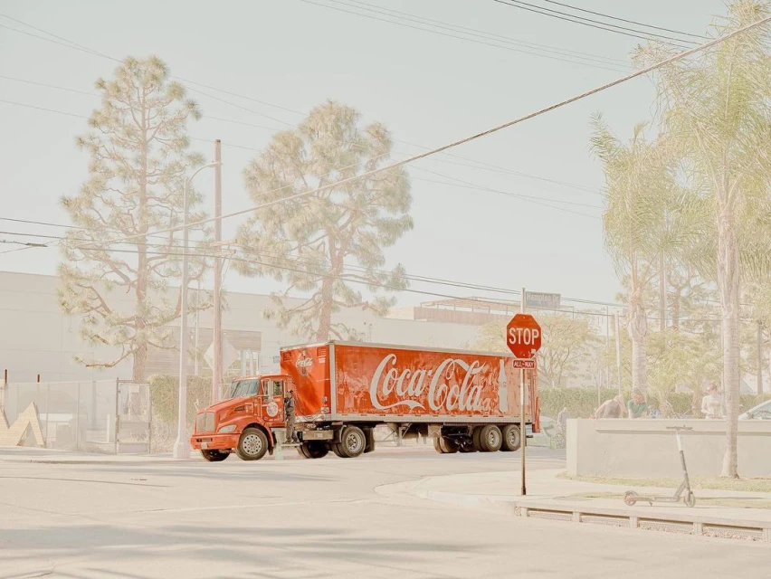 Red Coca-Cola delivery truck on urban street corner with faded light, trees, buildings, stop sign, and scooter in background.
