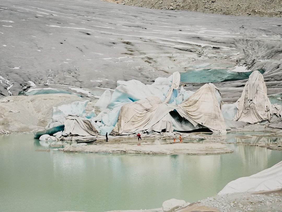 Melting glacier with a group of people in the foreground on a bright day, covered in protective white fabric, with glacier and light green water.