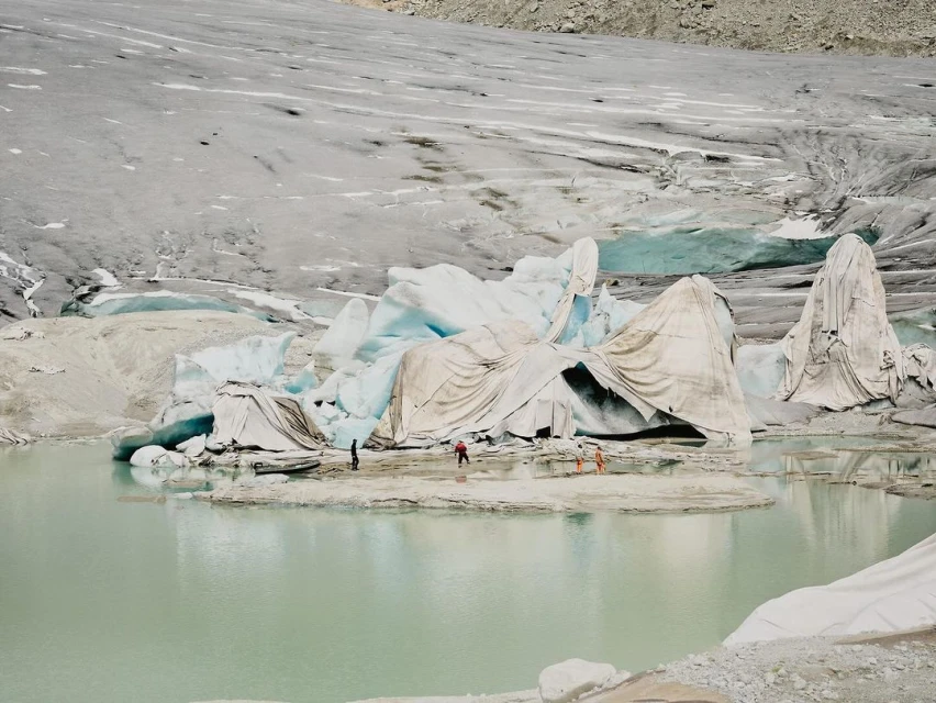 Melting glacier with a group of people in the foreground on a bright day, covered in protective white fabric, with glacier and light green water.