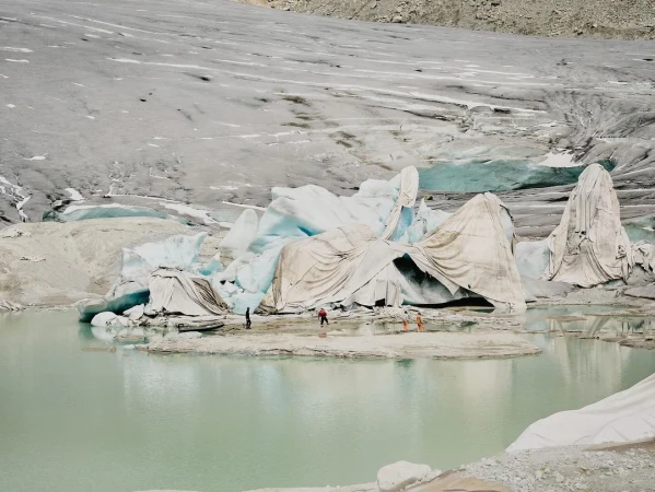 Melting glacier with a group of people in the foreground on a bright day, covered in protective white fabric, with glacier and light green water.