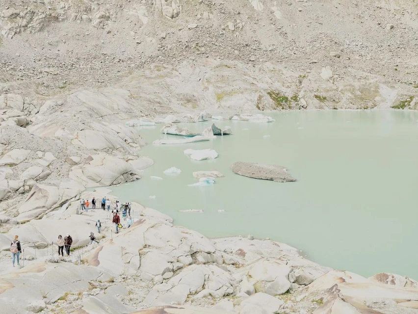 A group of people is near a glacial lake with icebergs on a sunny day. The landscape consists of rocky terrain and a pale blue-green lake.