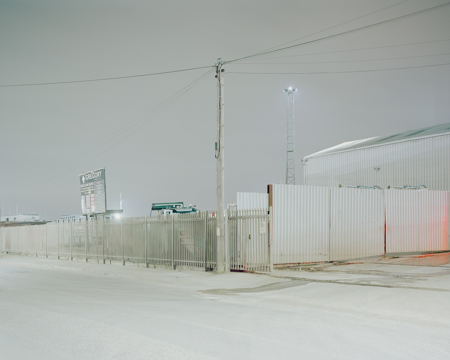 Industrial landscape with a chain-link fence, corrugated metal building, and utility pole under a cloudy sky, creating a stark, grey scene.
