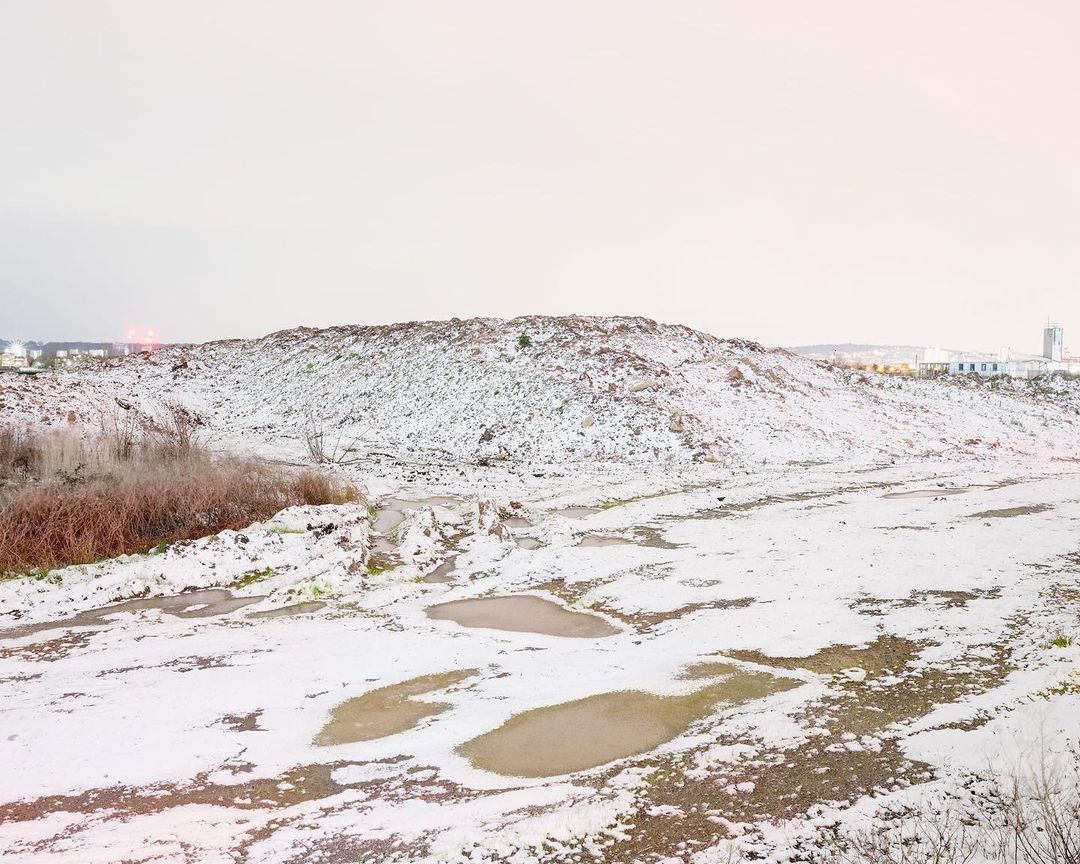 Winter landscape with snow-covered hill and muddy ground with puddles under a cloudy sky, buildings faintly visible in distance.