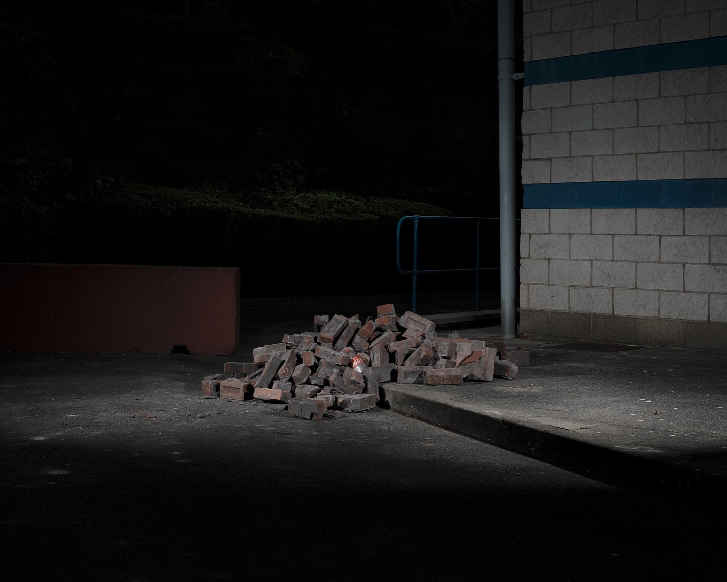 A pile of red bricks sits on a concrete surface next to a brick wall under dim lighting, creating a simple and somewhat mysterious outdoor scene.