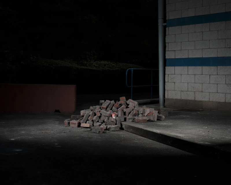 A pile of red bricks sits on a concrete surface next to a brick wall under dim lighting, creating a simple and somewhat mysterious outdoor scene.