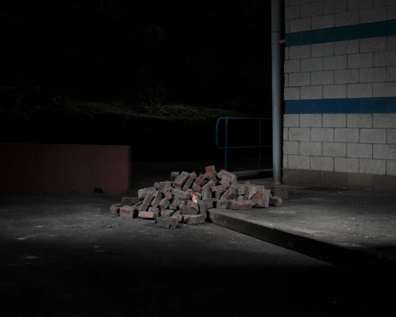 A pile of red bricks sits on a concrete surface next to a brick wall under dim lighting, creating a simple and somewhat mysterious outdoor scene.