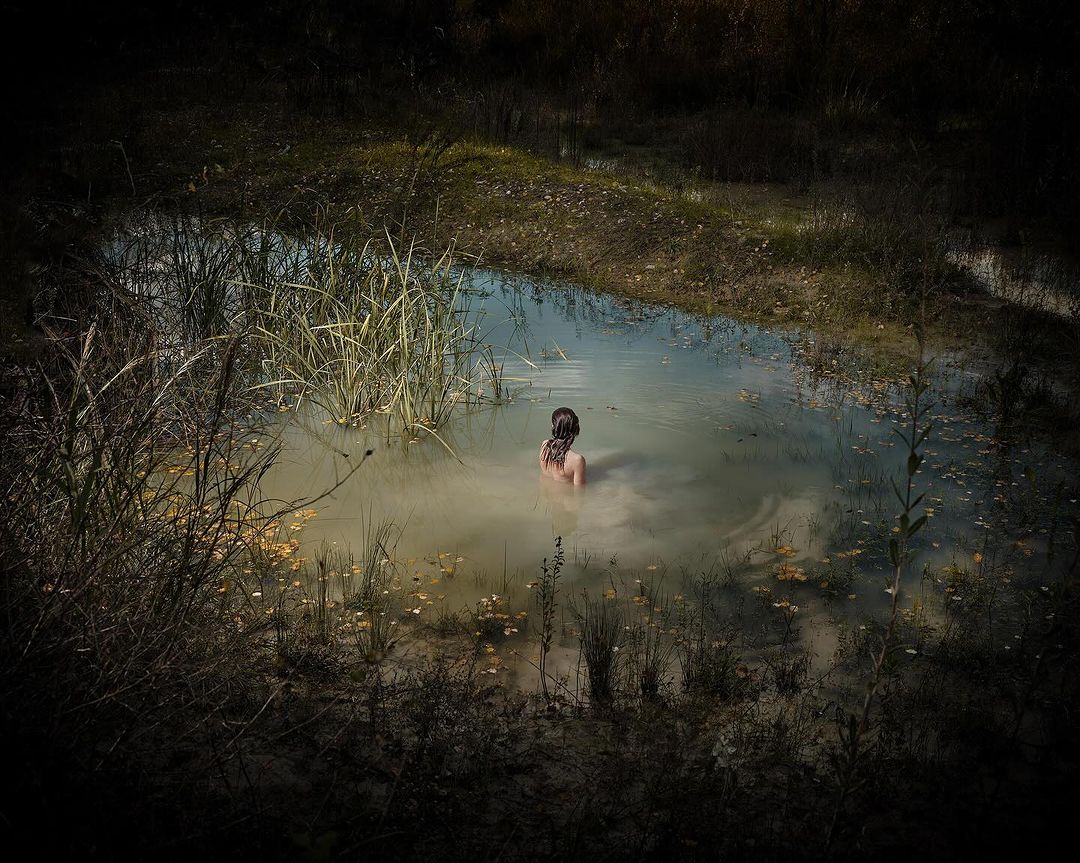 A young woman stands waist-deep in a murky pond surrounded by tall grasses and dark foliage in a moody, painterly outdoor scene.