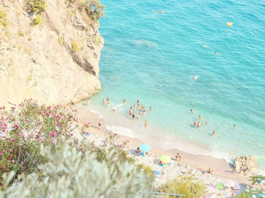 High-angle shot of a sandy beach with people swimming and relaxing in the ocean, beside a rocky cliff in bright daylight.