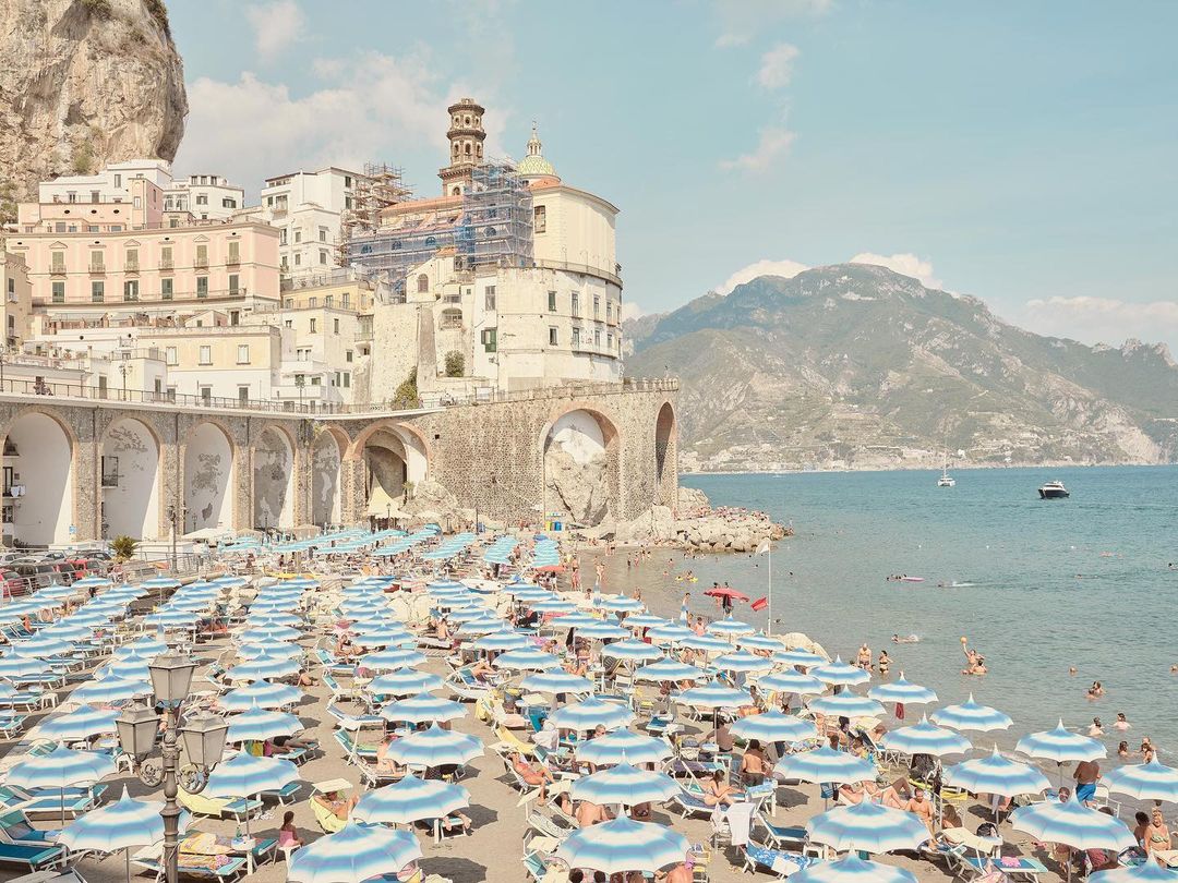 Panoramic beach view featuring blue umbrellas, buildings, and mountains in Amalfi, Italy, on a bright, sunny day, perfect for travel.