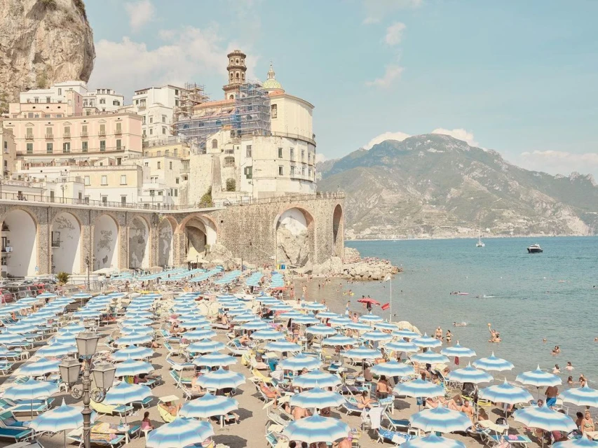 Panoramic beach view featuring blue umbrellas, buildings, and mountains in Amalfi, Italy, on a bright, sunny day, perfect for travel.