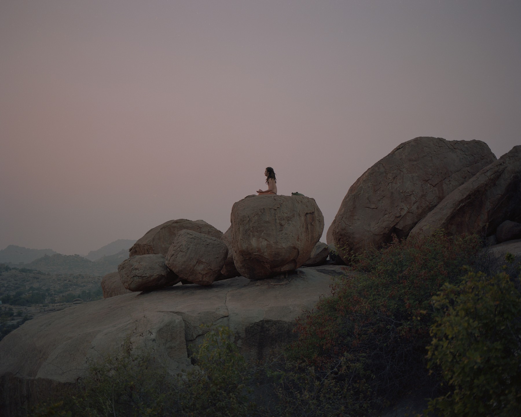 Woman sits in lotus position on large rocks with mountain backdrop at dusk. Relaxing nature scene, wellness, solitude, dusky natural light.