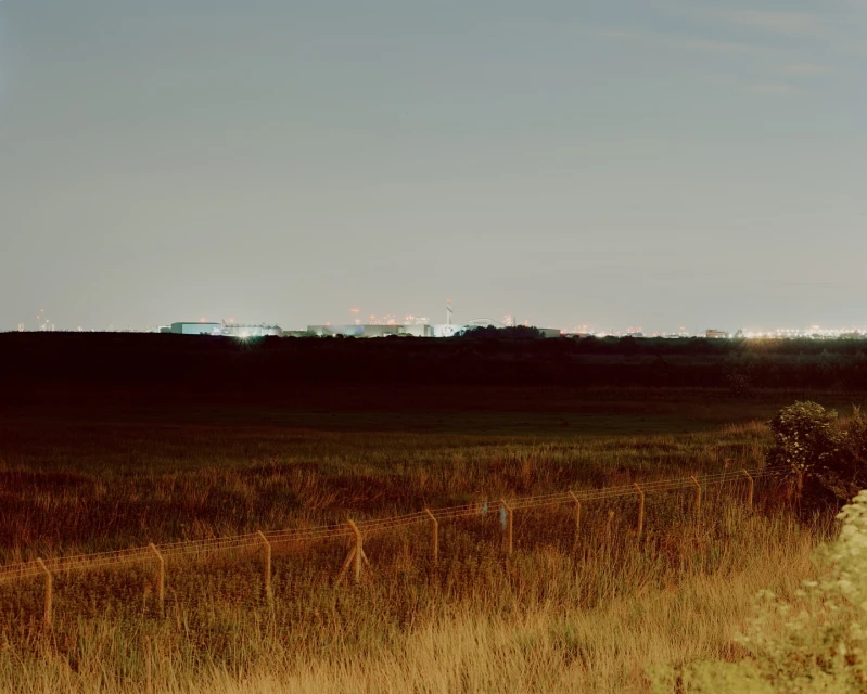 Field with sparse grass and a chain link fence in the foreground with an industrial area and sky in the background.