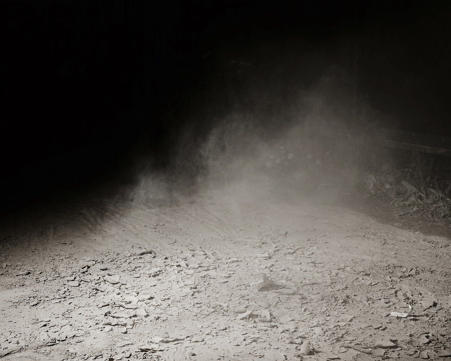 A dimly lit dirt road, dust particles suspended in air, contrasting with the dark surroundings. Low light creates shadows.
