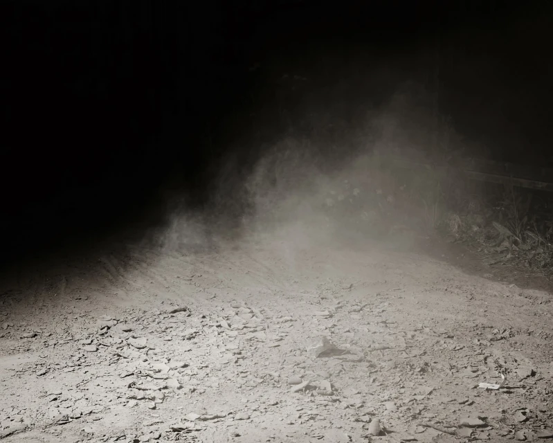 A dimly lit dirt road, dust particles suspended in air, contrasting with the dark surroundings. Low light creates shadows.