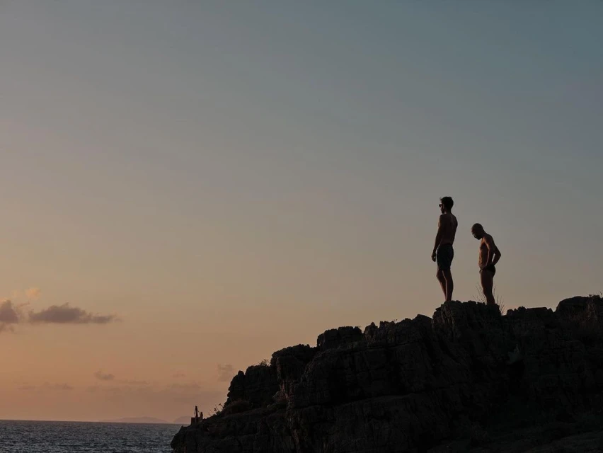 Two adult males silhouetted against a sky at dusk stand on a rocky outcrop overlooking the ocean in a lifestyle outdoor shot.