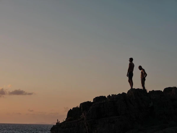 Two adult males silhouetted against a sky at dusk stand on a rocky outcrop overlooking the ocean in a lifestyle outdoor shot.