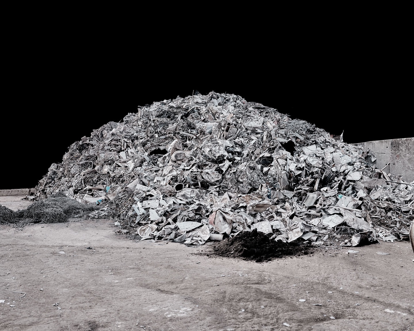Large pile of crushed and compacted silver metal scrap, with a flat, grey, concrete surface under it, and a dark background.