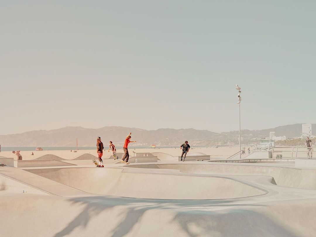 A group of skateboarders and scooter riders perform tricks in a concrete skatepark on a sunny beach in an outdoor recreation scene.